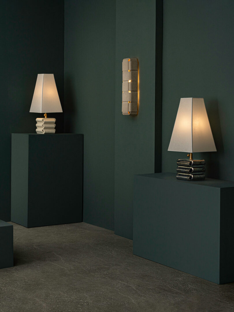Two table lamps with stacked stone bases and white shades—reminiscent of Virignia Sin lighting—sit on dark green pedestals; a matching wall sconce is mounted between them on a dark green wall