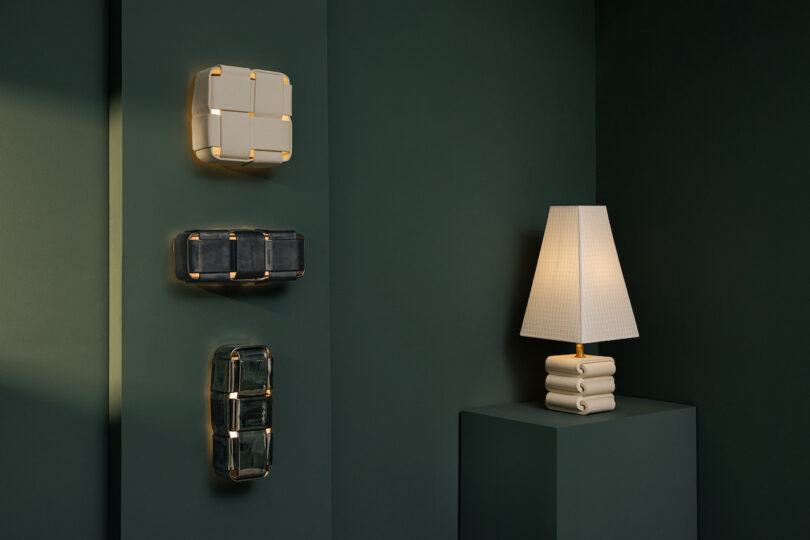 A cream table lamp with a pleated shade sits on a pedestal beside three geometric Virginia Sin lighting wall sconces mounted on a dark green wall