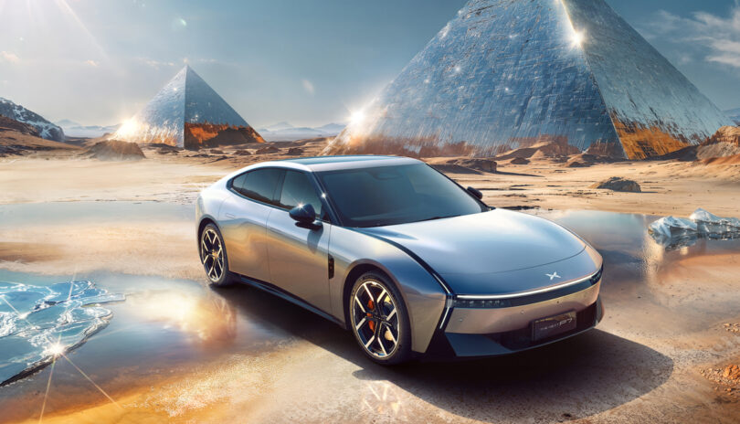 A sleek silver XPeng P7 electric car is parked on a reflective desert surface with futuristic glass pyramids in the background under a clear sky.