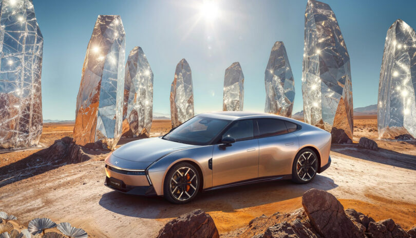 A silver XPeng P7 electric car is parked on a desert landscape, surrounded by large, reflective crystal structures under a bright sun.