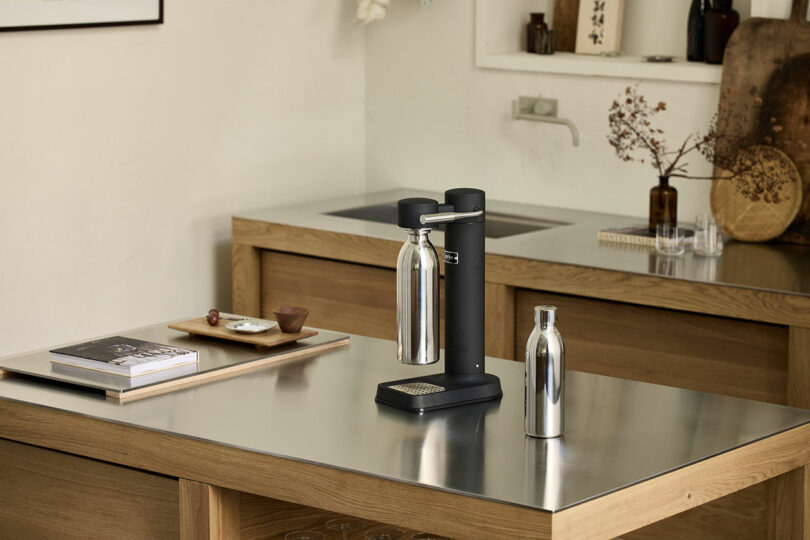 A modern kitchen with a sleek soda maker and a To-Go Bottle Aarke on a reflective countertop, surrounded by minimalist decor and wooden cabinetry.