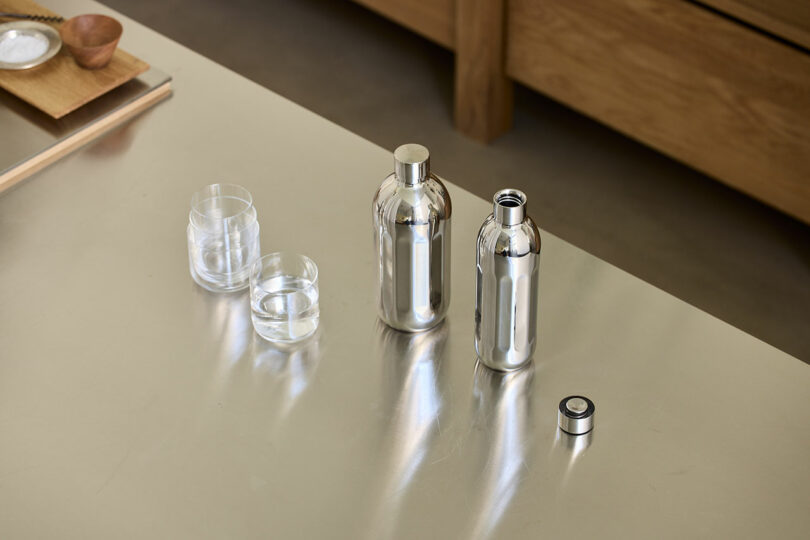Two stainless steel To-Go Bottles Aarke and two clear glasses filled with water sit on a reflective metallic surface; one bottle has its cap removed.