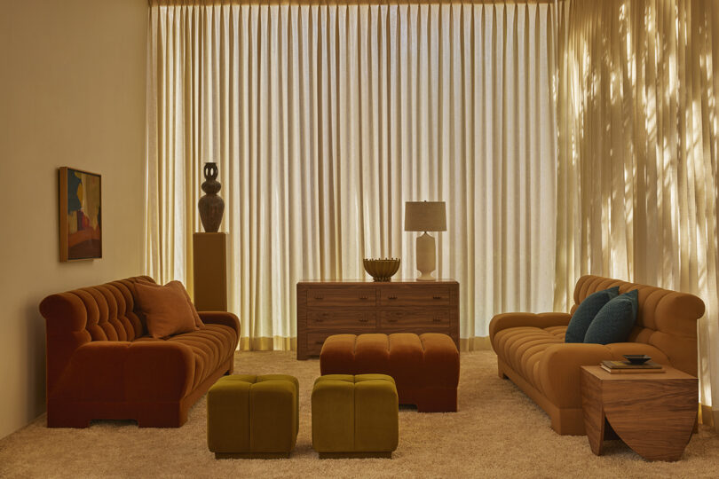A living room with two rust-colored sofas, a wooden sideboard, ottomans, a lamp, and a painting, with sheer curtains letting in natural light.