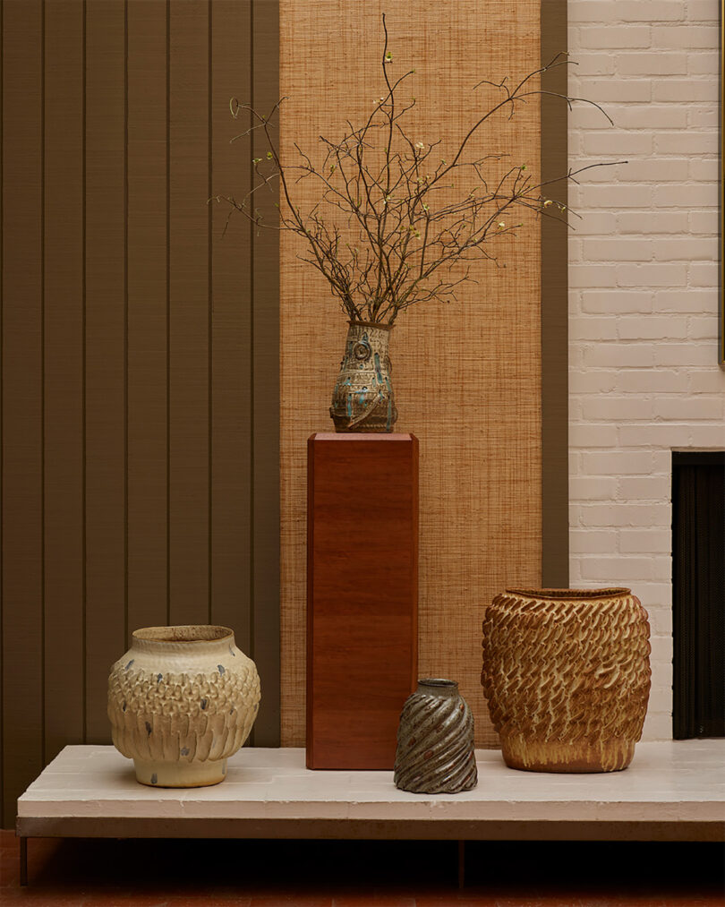 Four textured ceramic vases displayed on a marble surface; one vase with branches is elevated on a wooden pedestal against a neutral, modern wall background.