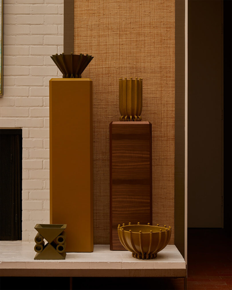 Four sculptural ceramic vessels in earthy tones are displayed on two tall pedestals and a white surface, set against a backdrop of textured wall panels and white brick.