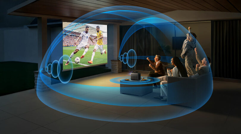 Four people sit outside at night watching a soccer match on a Nebula X1’s large screen, with illustrated sound waves surrounding them to depict immersive audio.