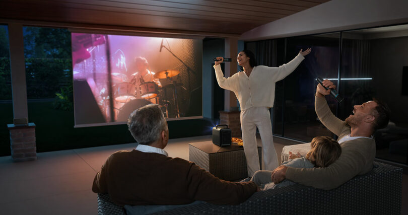 Four people enjoy a karaoke night on the patio; two sing with microphones while a concert video streams on the Nebula X1 projector, displayed on a large outdoor screen in the background.
