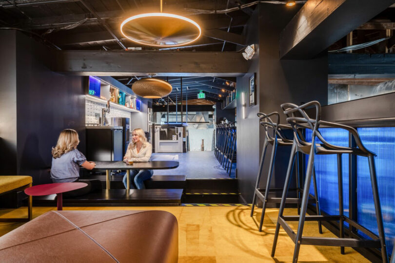 Two people sit and talk at a table in a modern, dimly lit lounge area with bar seating, overhead lighting, and contemporary decor.