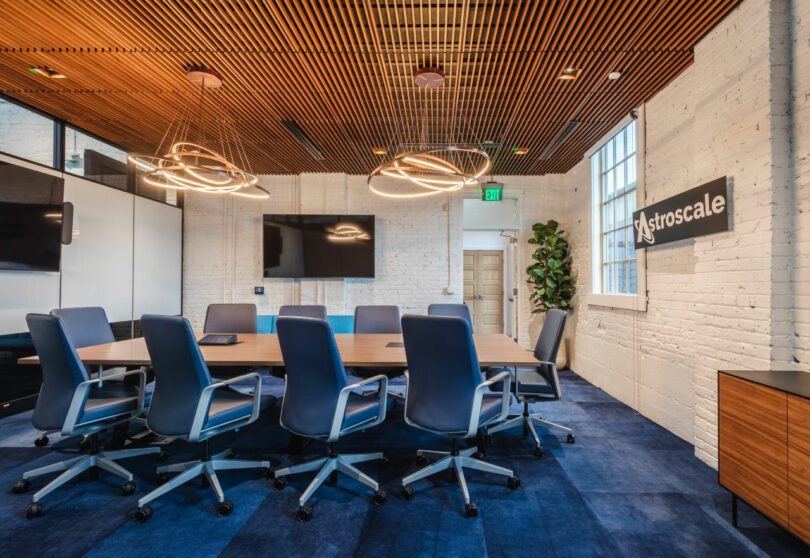 Modern conference room with a long table, blue chairs, wall-mounted screens, geometric light fixtures, and a sign reading "Astroscale" on a white brick wall.