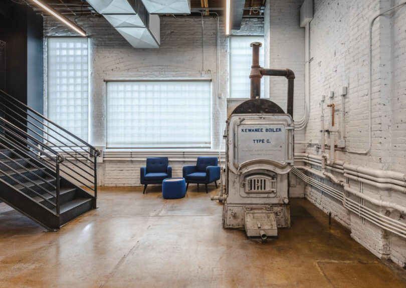 Industrial-style room with an old Kewanee Boiler, two blue chairs, an ottoman, exposed pipes, and metal stairs against white brick walls and large windows.