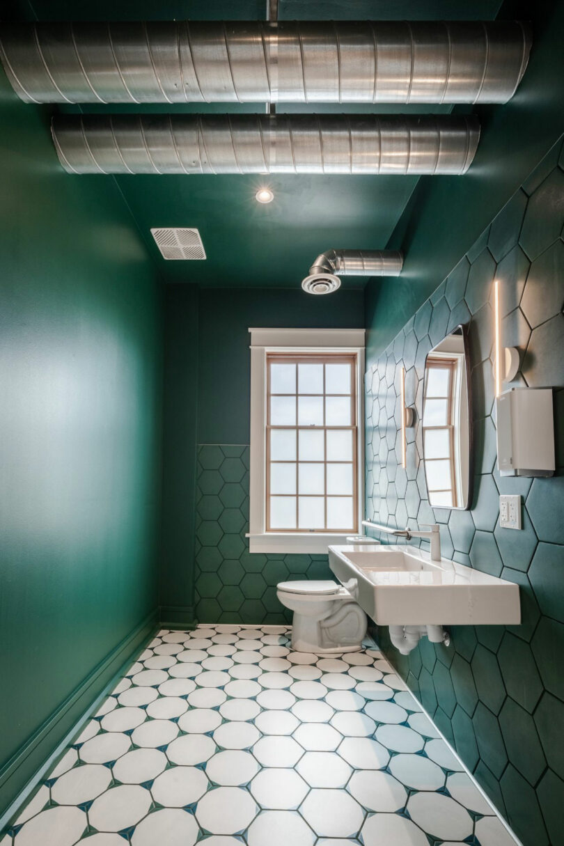 A modern bathroom with green hexagonal wall tiles, a large window, exposed metal ductwork, a wall-mounted sink, and a toilet on a white hexagon-tiled floor.