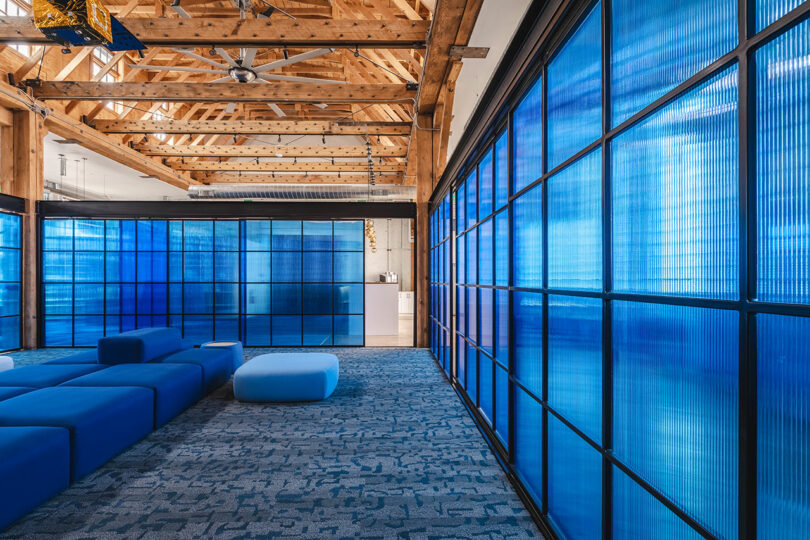 Modern office lounge with blue modular seating, patterned carpet, exposed wooden beams, and large blue-tinted translucent wall panels—a sleek space that reflects Astroscale’s forward-thinking design ethos.
