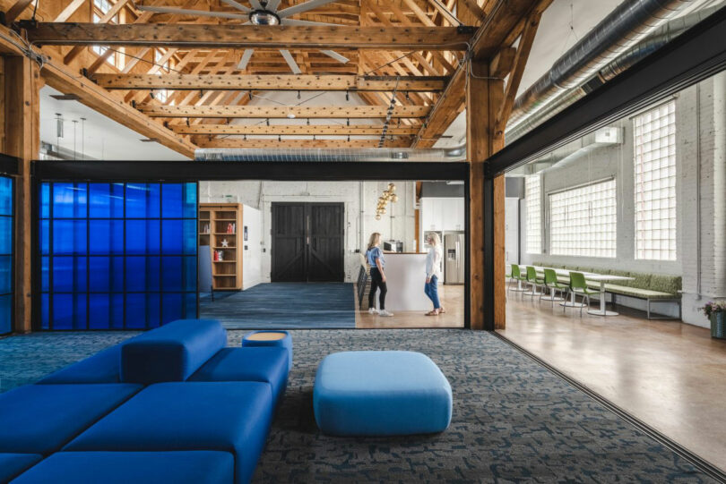 Modern open office space with exposed wooden beams, blue seating in the foreground, and two people conversing near a white reception area in the background.