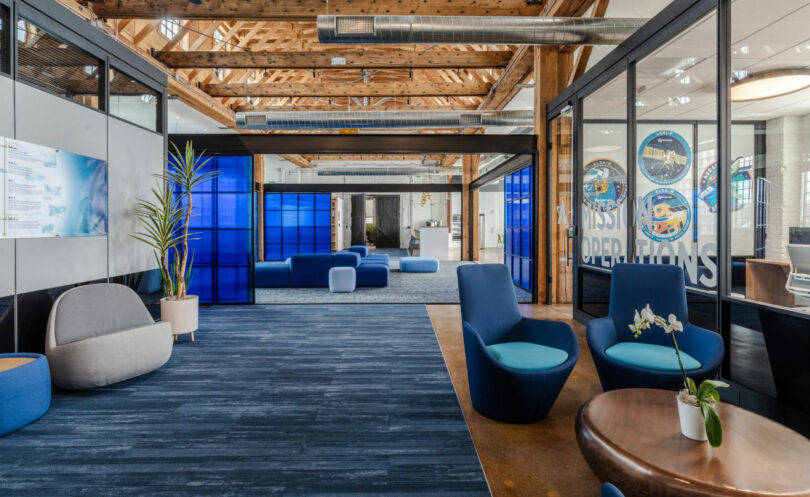 Modern office lounge area with blue and gray chairs, a small plant on a round table, exposed wooden beams, and blue partition walls.
