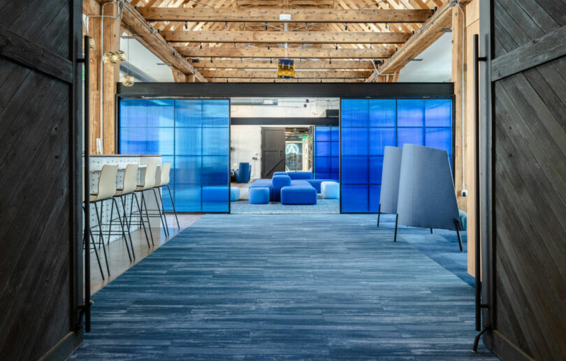 Modern office space with exposed wooden beams, blue carpet, high chairs at a counter, and blue-tinted glass partitions dividing seating areas.