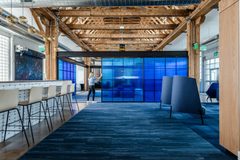 A person stands in a modern open office space with exposed beams, blue carpet, bar seating, and large blue-tinted glass panels.
