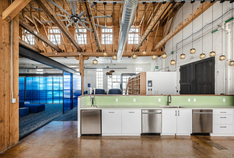A modern office kitchen with green tile backsplash, white cabinets, and stainless steel appliances by Astroscale, adjacent to an open workspace with exposed wood beams and large windows.