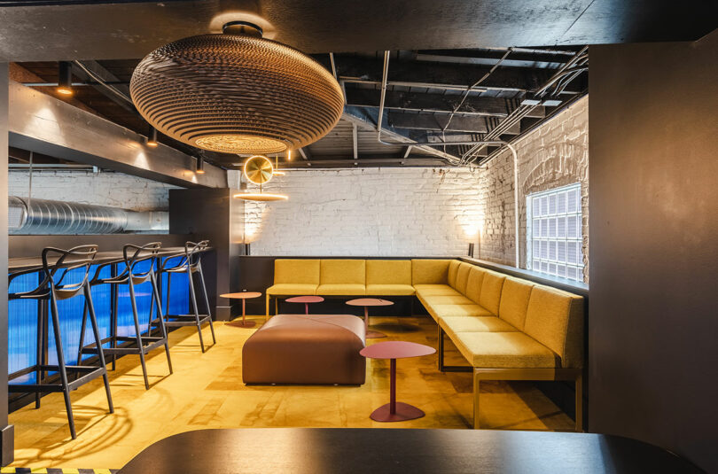 Modern lounge area with yellow sectional seating, small round tables, bar stools, and industrial-style lighting in a room with exposed brick walls and ceiling pipes—an inviting space inspired by Astroscale’s innovative design ethos.
