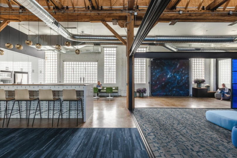 Modern office space with exposed beams, a kitchen area with bar stools, lounge seating, and large windows letting in natural light. Two people are seated in different areas.