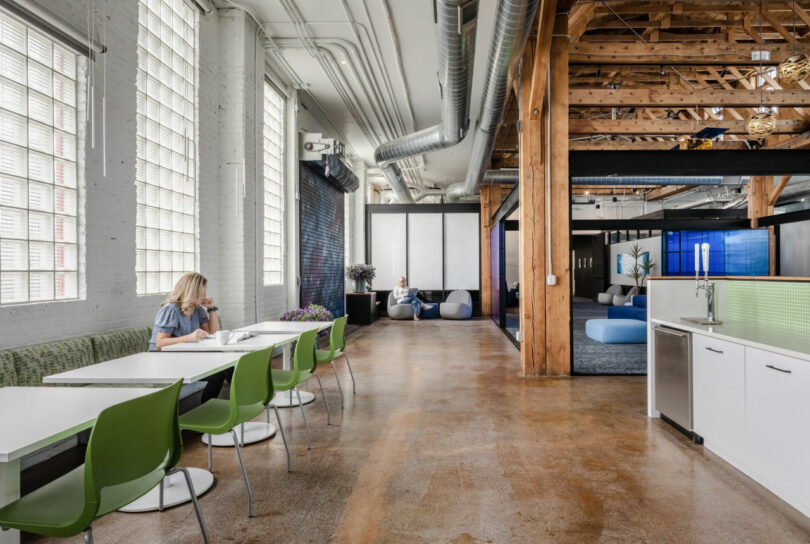 A woman sits at a table in a modern office with green chairs, exposed wooden beams, large windows, and an open workspace in the background.