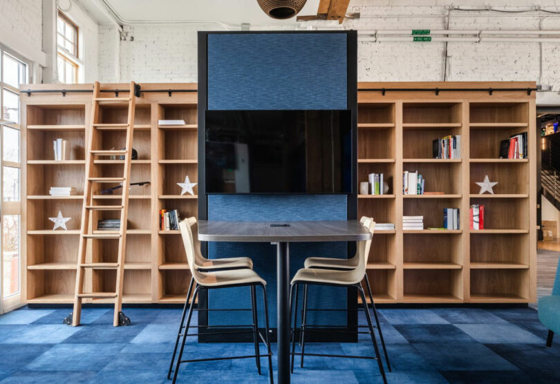 A modern workspace with a tall table and chairs in front of built-in wooden bookshelves, a mounted screen, and a rolling ladder against a white brick wall.