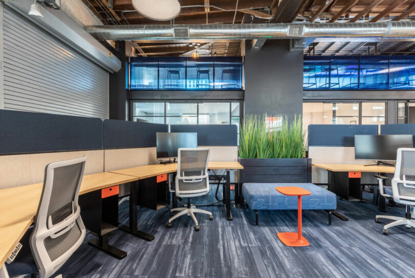 Modern open-plan office with desks, chairs, monitors, blue partition panels, and a planter divider. Exposed ceiling, overhead ducts, and glass walls are visible in the background.