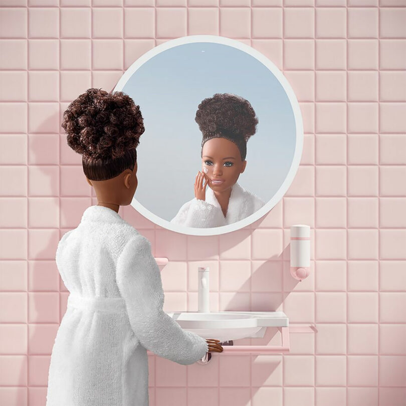 A Barbie x HEWI doll with curly hair in a white robe stands at a sink, gazing at herself in a round mirror on a pink tiled wall.