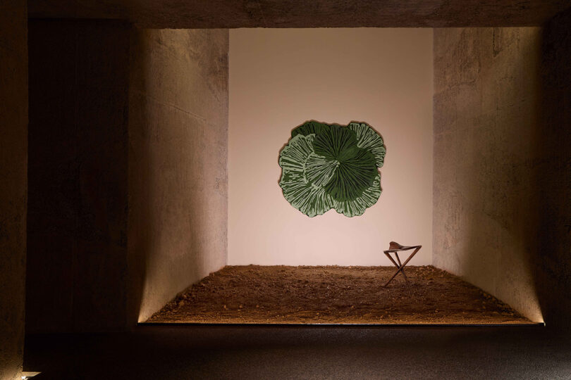 A minimalist room with textured walls, a dirt floor, a green leaf-shaped wall art, and a small wooden stool holding a ceramic bowl.
