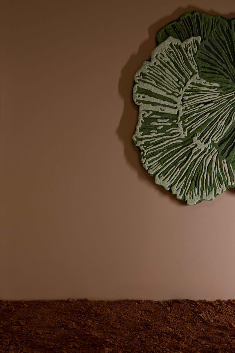 A large, green, leaf-shaped textile wall decoration hangs on a plain brown wall above a rough-textured brown floor.