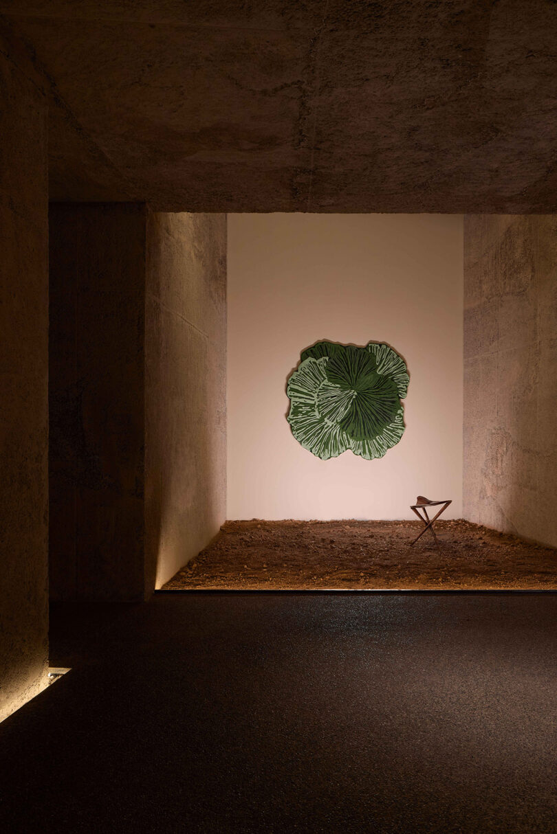 A minimalist room features a large green leaf wall art, a simple folding stool, and a dirt-covered floor under soft, warm lighting.