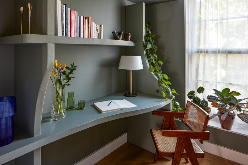 A modern home office corner with a built-in desk, books, plants, a lamp, an open notebook, and a wooden chair by a window with sheer curtains.