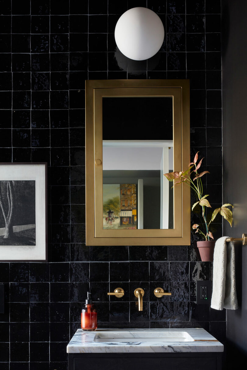 A bathroom with glossy black tile walls, a gold-framed mirror, a marble sink, a round wall light, a towel on a bar, and soap and a potted plant on the counter.