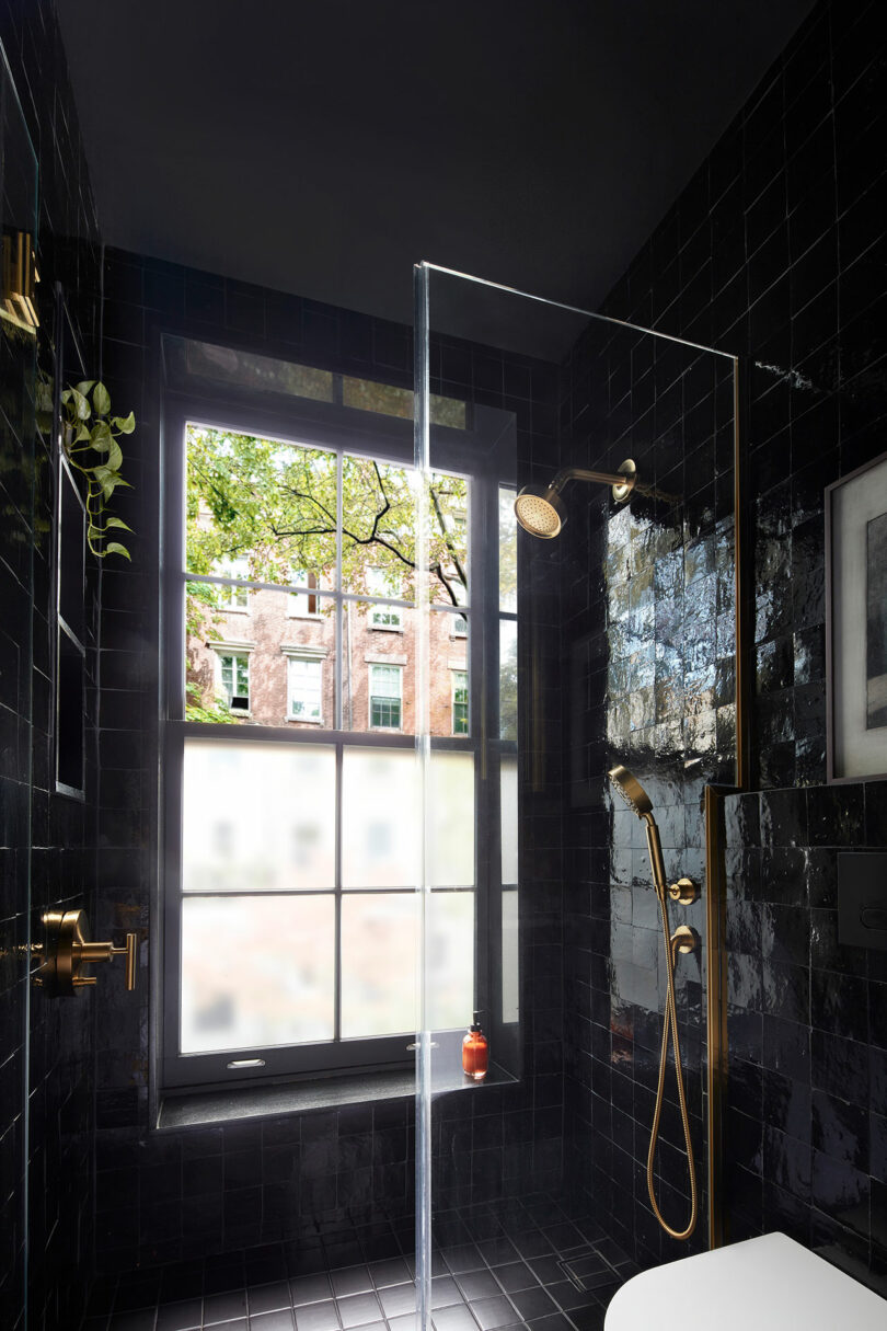 Modern shower with glossy black tiles, gold fixtures, a glass door, and a large window overlooking trees and a brick building outside.