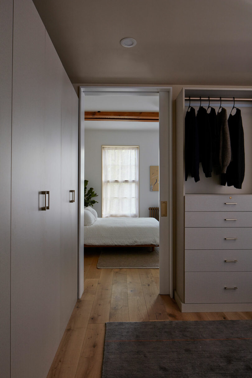 A view from a closet with hanging clothes and drawers into a bedroom with a bed, a window with sheer curtains, and wooden flooring.