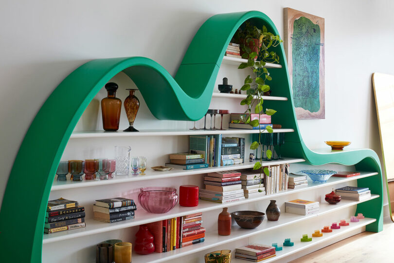 A green, wave-shaped bookshelf holds books, glassware, vases, a potted plant, and decorative objects against a white wall with framed art.