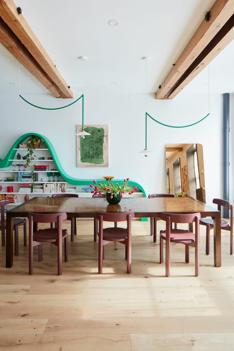 Modern dining room with a wooden table, eight mauve chairs, exposed ceiling beams, light wood floors, a vase of red tulips, and green wave-like shelving on a white wall.