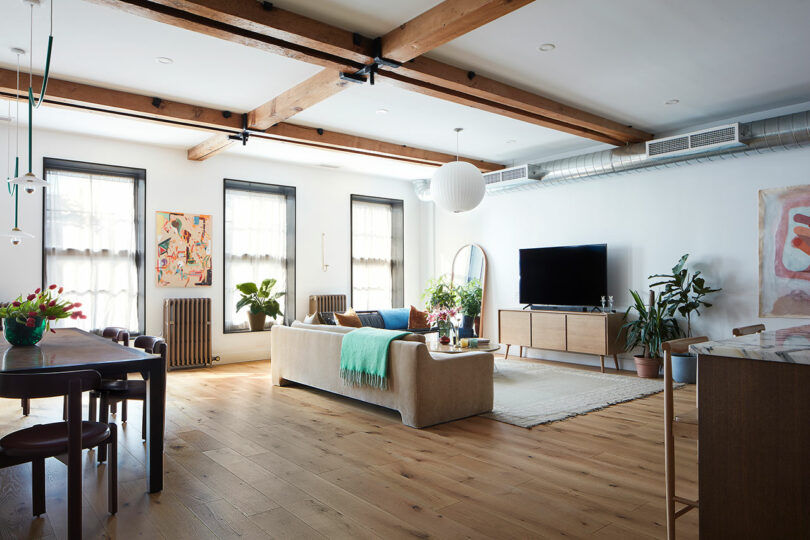 A modern living room with wooden beams, large windows, light wood floors, a beige sofa, dining table, TV, houseplants, and contemporary art on white walls.