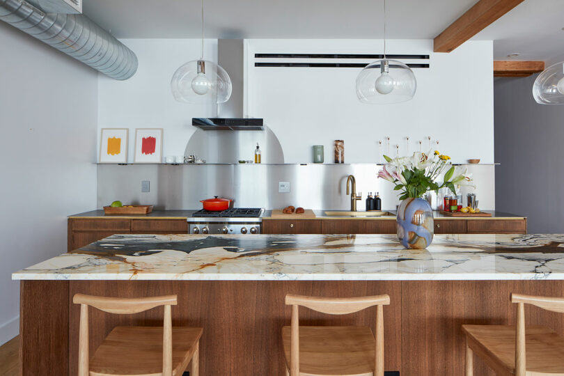 Modern kitchen with a marble island, wooden chairs, stainless steel appliances, pendant lights, and a vase of flowers on the counter.