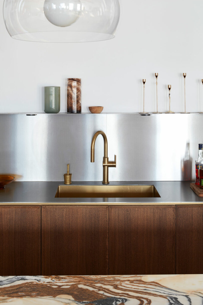 Modern kitchen with a brass faucet and sink, wooden cabinets, stainless steel backsplash, and decorative items on the countertop and shelf.