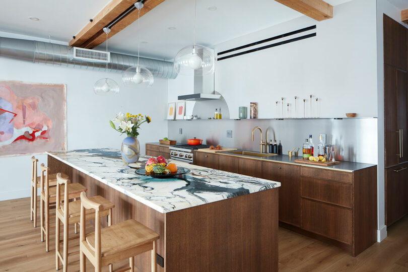 Modern kitchen with wooden cabinets, a marble island, and wooden bar stools. Pendant lights hang from the ceiling, and a colorful painting is on the wall.