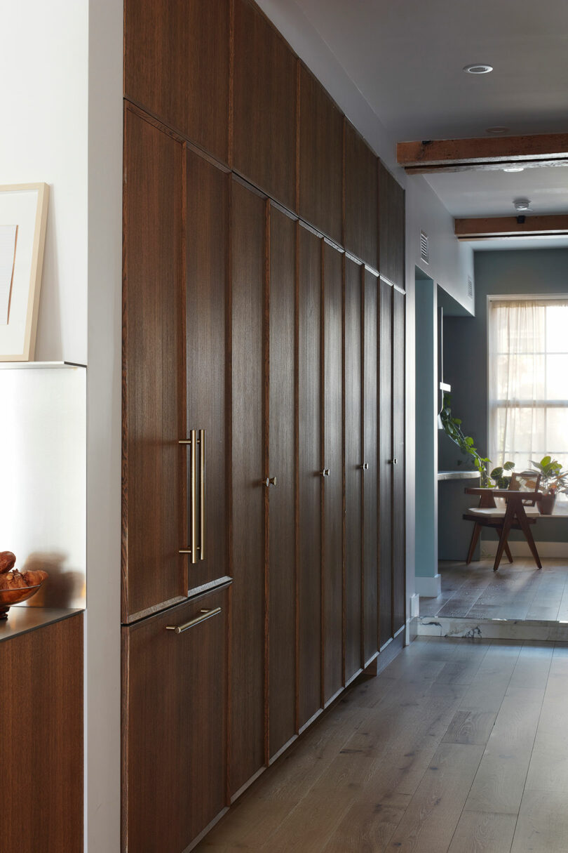 A hallway with floor-to-ceiling wooden cabinets on the left, light wood flooring, and a room with a dining table and chairs visible in the background.
