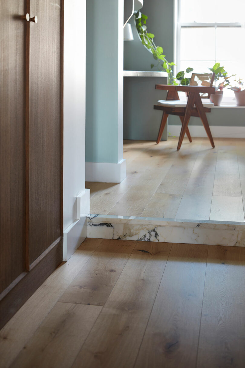 A room with light wood flooring, a step with a marble riser, a wooden desk and chair, potted plants, and sunlight coming through a window.