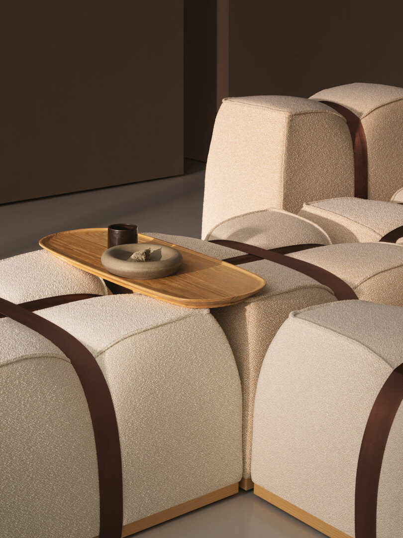 Beige modular seating by Mizetto with textured fabric and brown straps, topped with a wooden tray holding a ceramic dish and cup, set in a minimal, neutral-toned room.