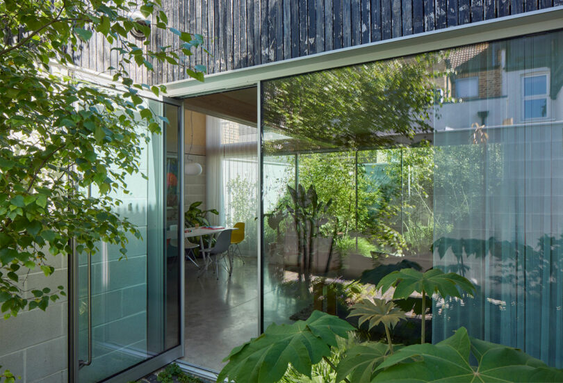 A modern glass sliding door opens to a dining area with chairs, surrounded by greenery and trees reflected in the glass.