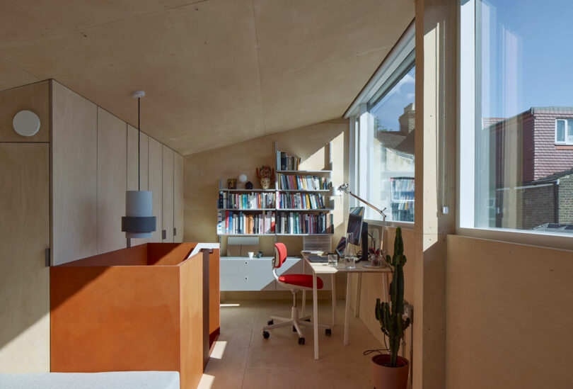 A modern home office with a built-in desk, red chair, bookshelf, potted cactus, and large windows letting in natural light.