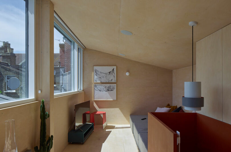 A minimalist loft room with sloped ceiling, large windows, light wood walls, a gray sofa, red chair, wall art, and a cactus near the window.