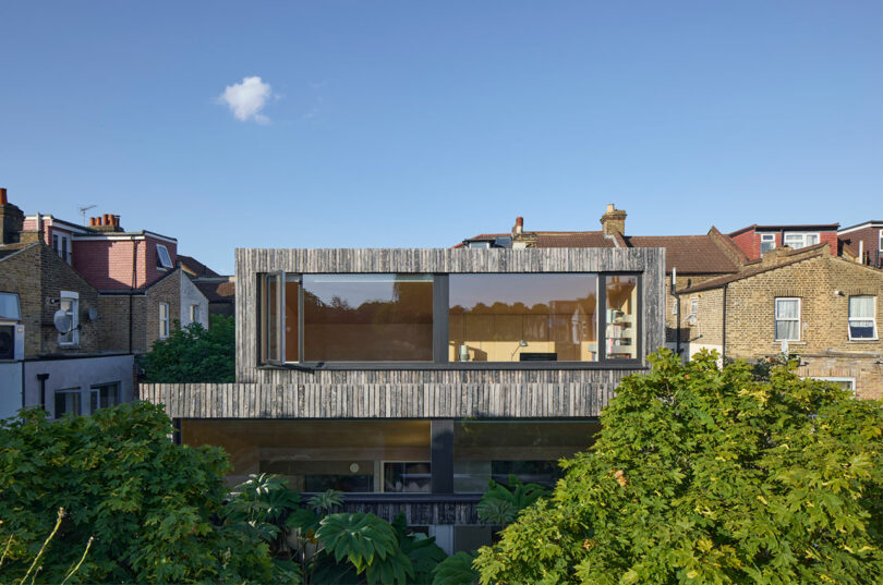 Modern house with a large horizontal window on the upper floor, surrounded by trees, set among traditional brick homes under a clear blue sky.
