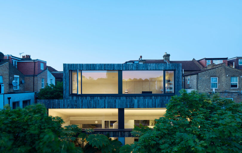 Modern two-story house with large horizontal windows and wooden exterior, surrounded by trees, set among traditional brick houses at dusk.
