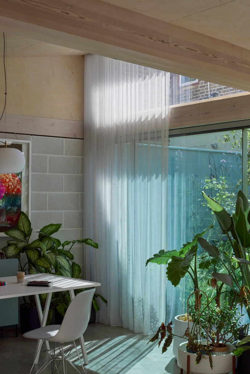 A bright room with large windows, sheer white curtains, potted plants, a white chair, and a table with a colorful hanging light fixture.