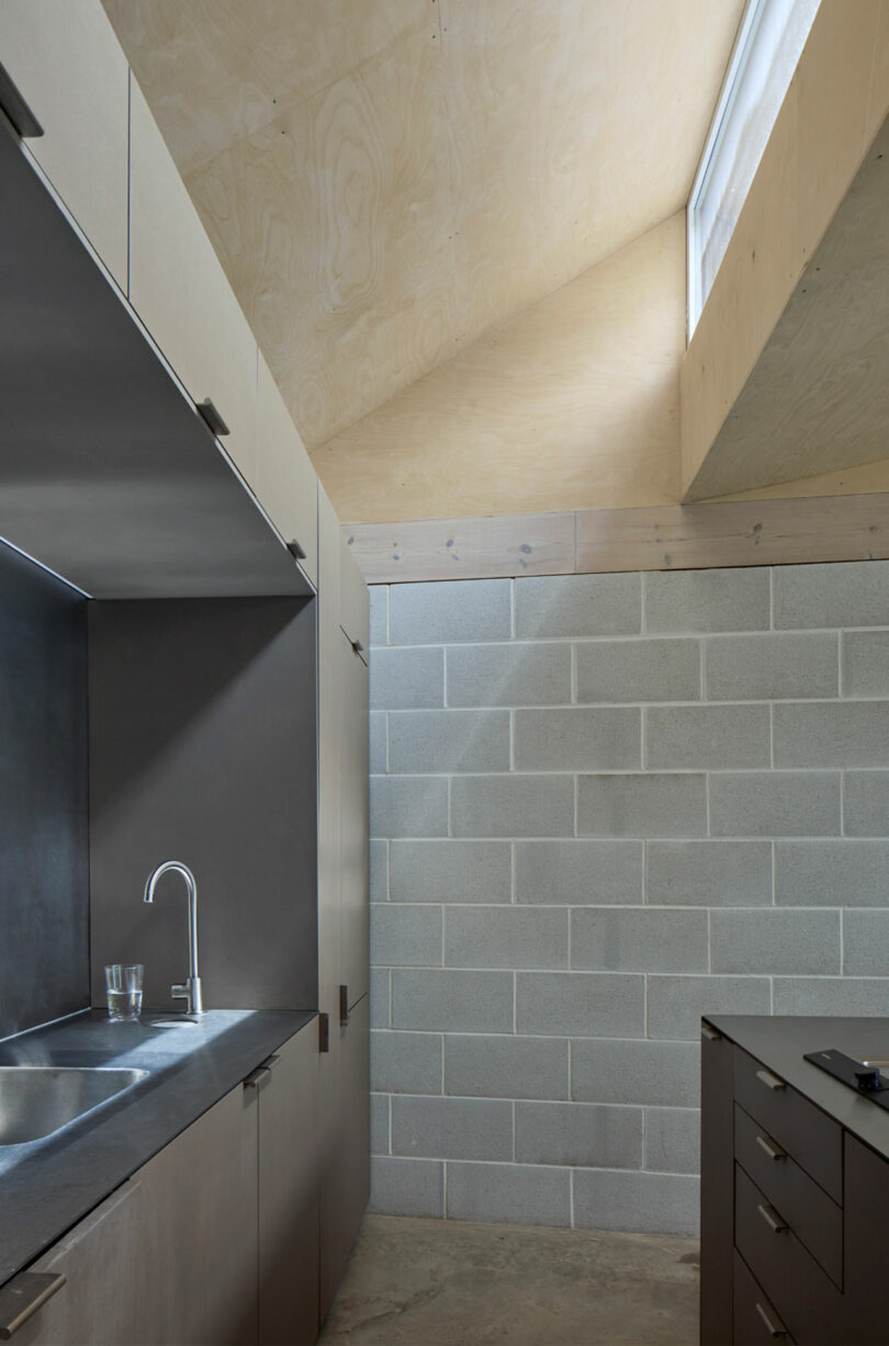 A modern kitchen with concrete block walls, light wood cabinets, a stainless steel sink, and a slanted ceiling with a high window letting in natural light.